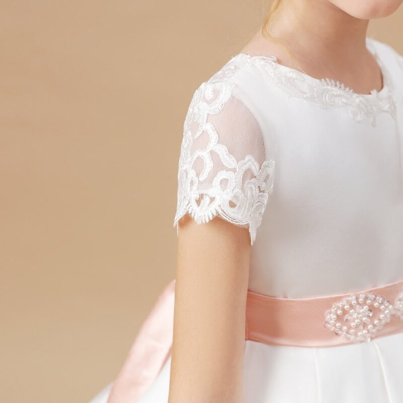 Close up of ivory satin flower girl dress featuring sheer embroidered lace sleeve, blush pink satin sash and pearl embellished waistband