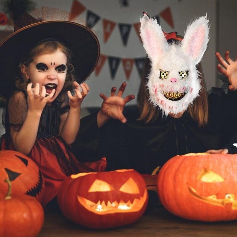Child wearing a white horror rabbit mask surrounded by Halloween pumpkins and decorations